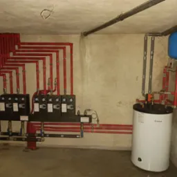 Złożony system instalacji grzewczej z rozdzielaczami, czerwonymi rurami i białym zasobnikiem Vaillant z niebieskim naczyniem przeponowym, zamontowany w piwnicy o surowych ścianach.