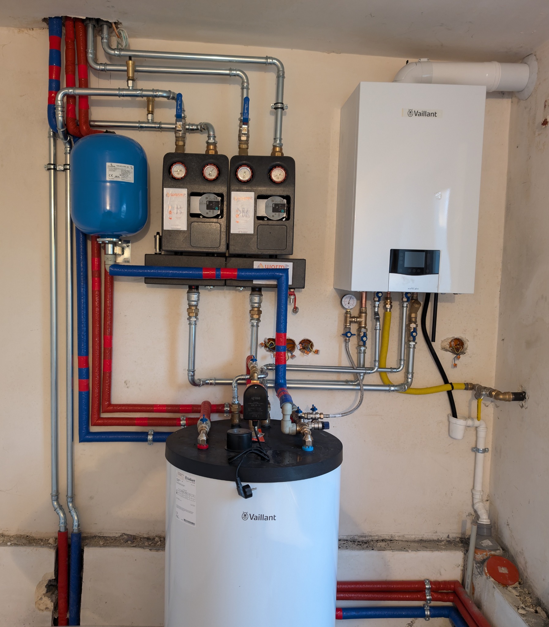 Zestaw urządzeń grzewczych Vaillant: piec gazowy, zasobnik ciepłej wody i rozdzielacze, z rurami w izolacji czerwono-niebieskiej, zamontowany przy ścianie w surowym pomieszczeniu.