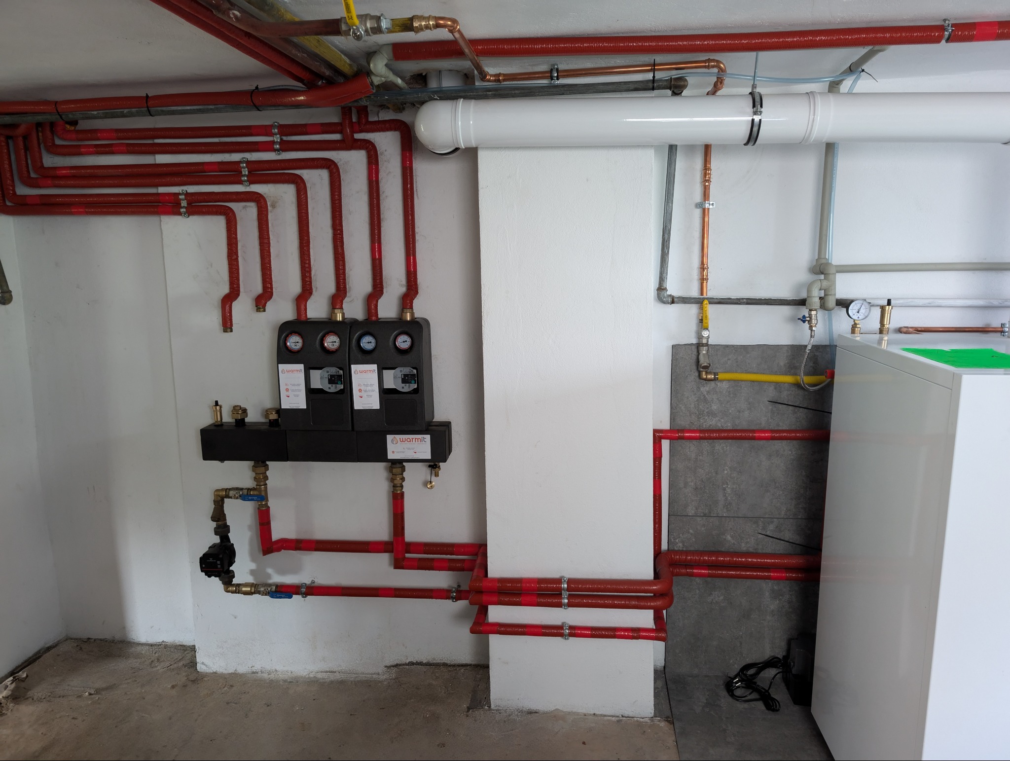 Rozbudowana instalacja grzewcza z czerwonymi rurami, czarnym rozdzielaczem Warmit z manometrami, białą rurą wentylacyjną i kotłem grzewczym w kotłowni.