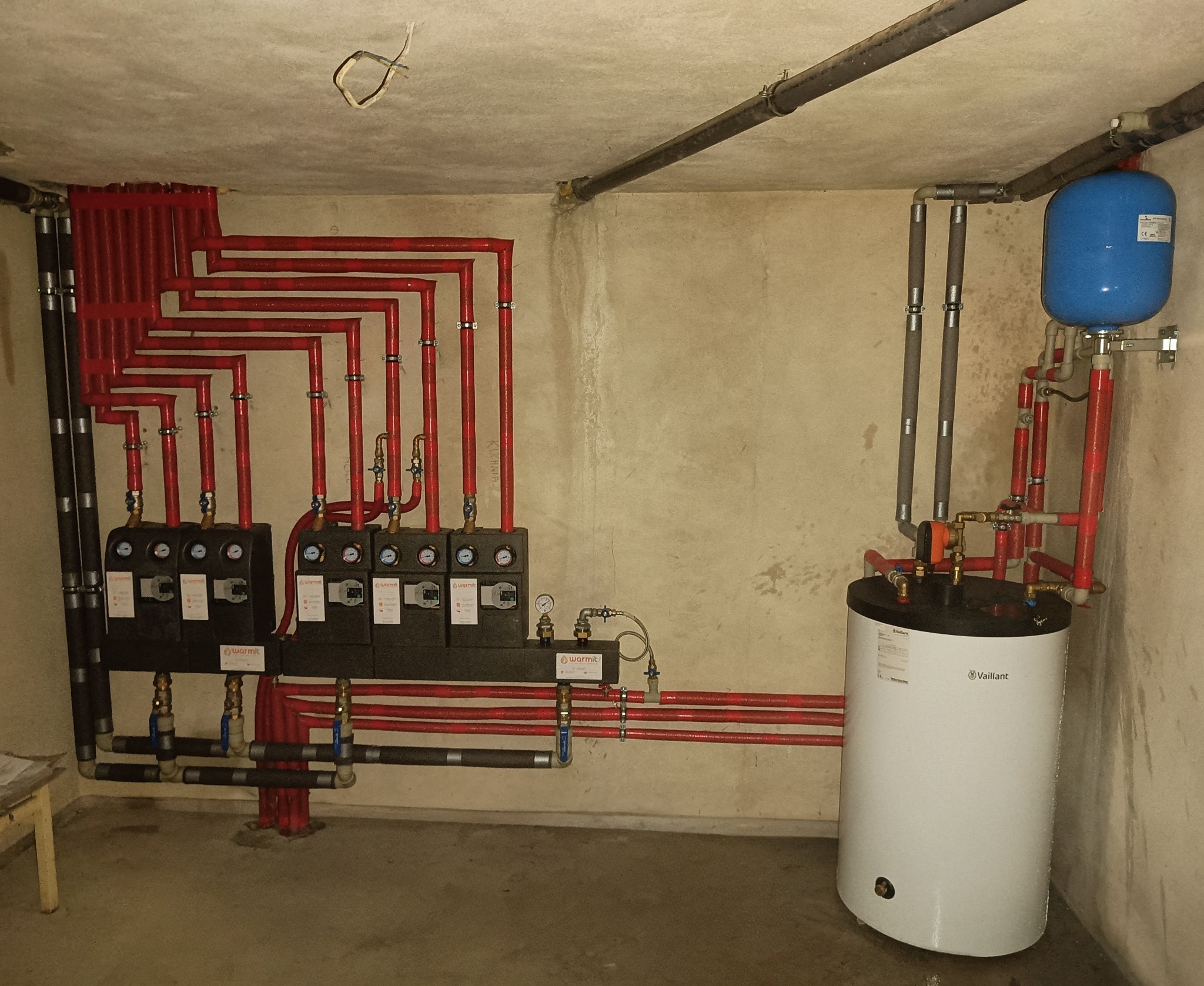 Złożony system instalacji grzewczej z rozdzielaczami, czerwonymi rurami i białym zasobnikiem Vaillant z niebieskim naczyniem przeponowym, zamontowany w piwnicy o surowych ścianach.