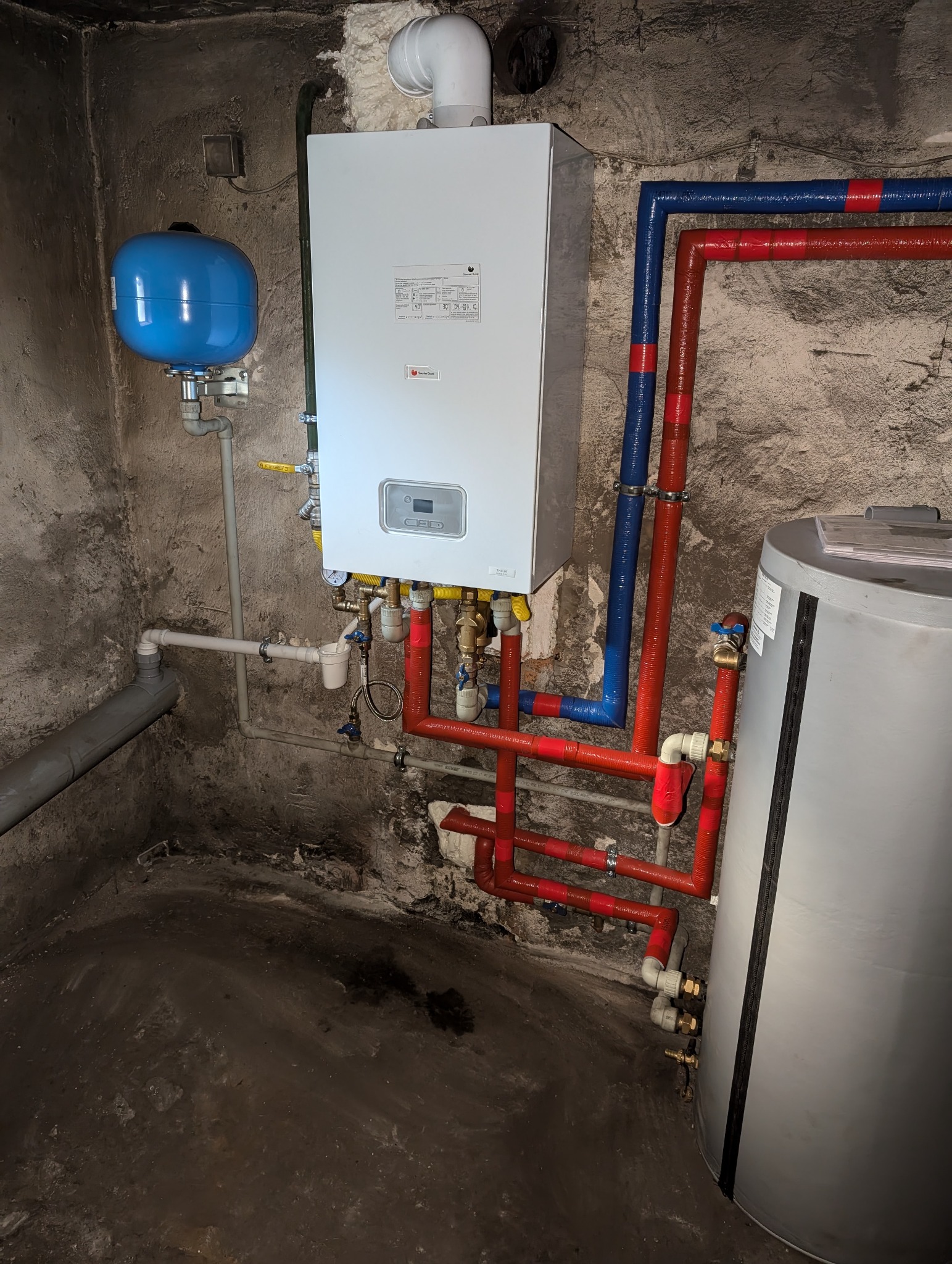 Nowoczesny piec gazowy wiszący z podłączonymi rurami w kolorach czerwonym i niebieskim, zamontowany na tle surowej, betonowej ściany w piwnicy.