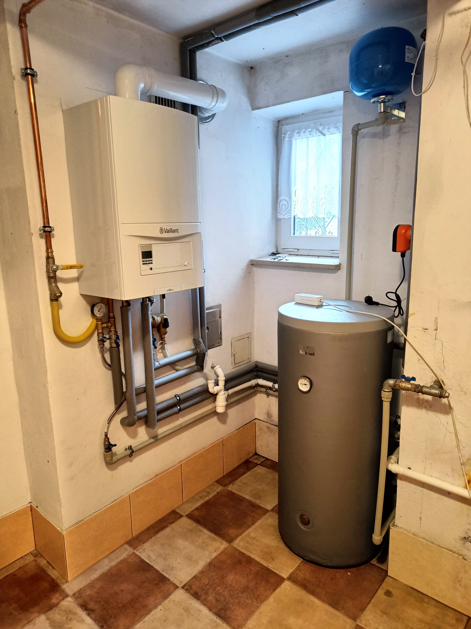 Biały piec gazowy Vaillant, szary zasobnik wody i niebieski zbiornik wyrównawczy w kotłowni z oknem i brązowo-beżową podłogą w kratkę.