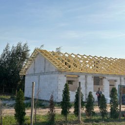 Klaudiusz Kończal - Wznoszenie konstrukcji dachu na budynku z białych bloczków, widoczna drewniana więźba dachowa, pracownik w tle i dostawczy samochód obok.