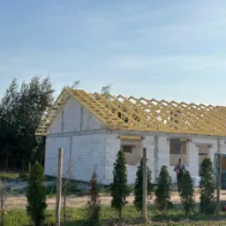 Wznoszenie konstrukcji dachu na budynku z białych bloczk&oacute;w, widoczna drewniana więźba dachowa, pracownik w tle i dostawczy samoch&oacute;d obok.