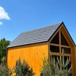 Nowoczesny dom z drewnianą elewacją i szarym dachem z dachówki, na tle błękitnego nieba. Widoczne detale wykończenia i roślinność w otoczeniu budynku.