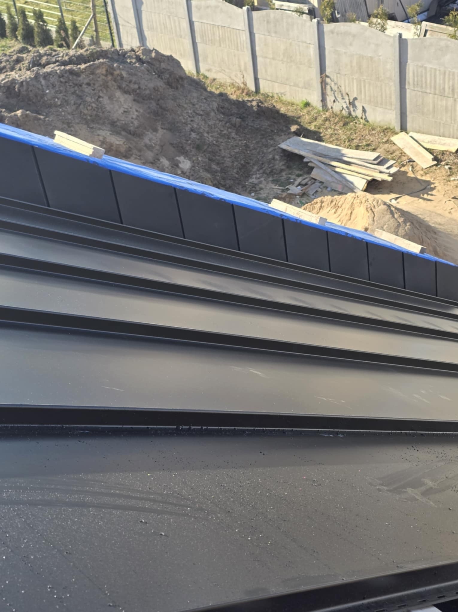 Nowe, czarne blaszane pokrycie dachowe z widocznymi łączeniami, zabezpieczone niebieską folią, w tle hałda ziemi i betonowy płot.