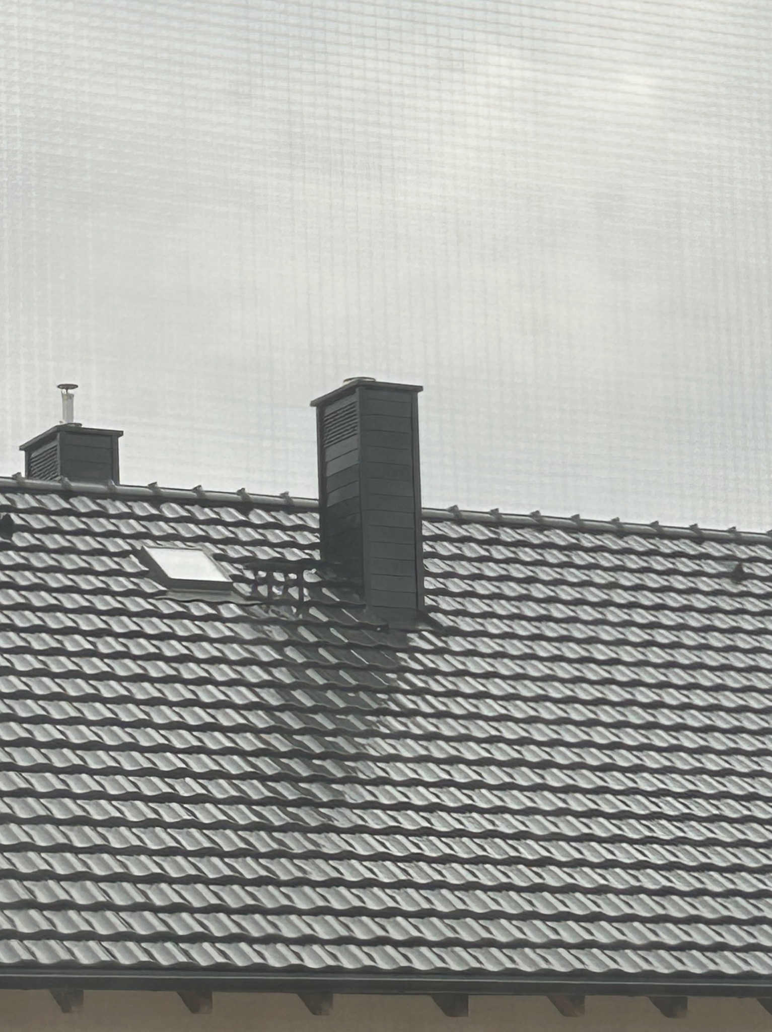 Czarny dach z dachówki karpiówki, dwa kominy z czarnej blachy, okno dachowe, widok na szary dzień.