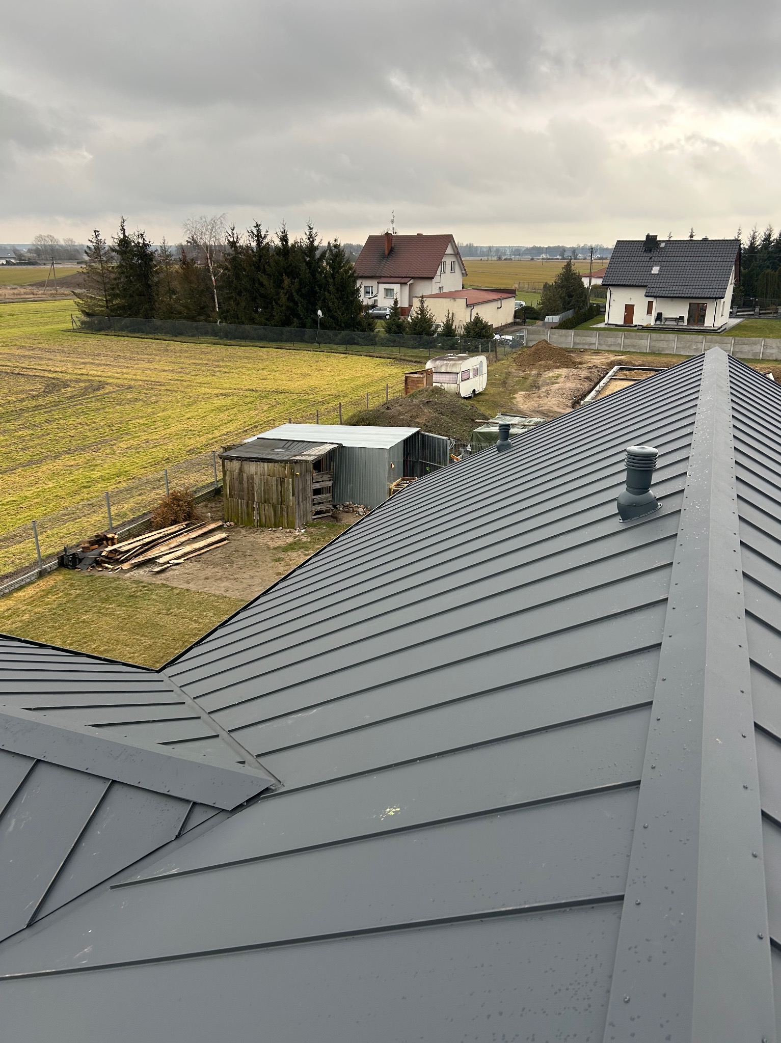 Nowy, szary dach z blachy na tle jesiennego krajobrazu wiejskiego z widocznymi kominkami wentylacyjnymi.