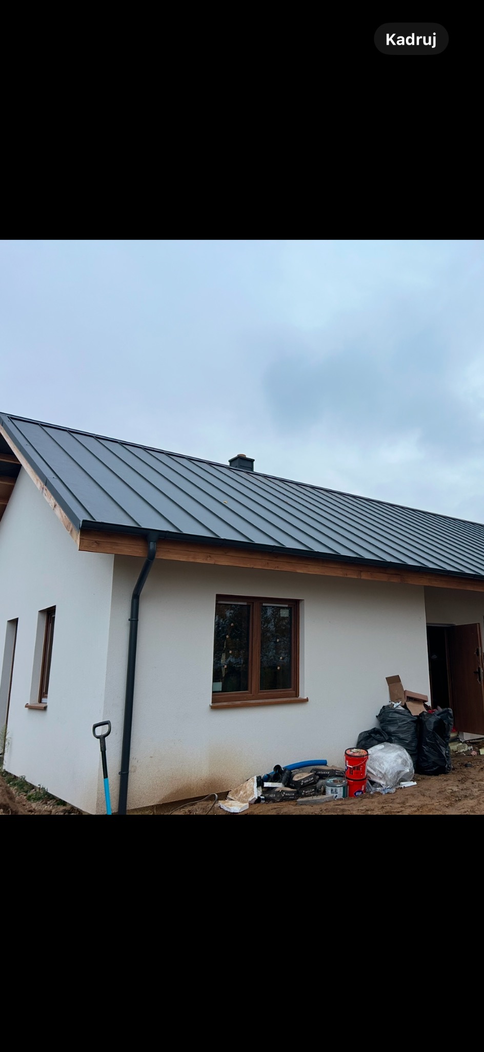 Nowy dom jednorodzinny z ciemnoszarą blachodachówką, brązowymi oknami i rynnami, widoczne ślady prac budowlanych wokół budynku.