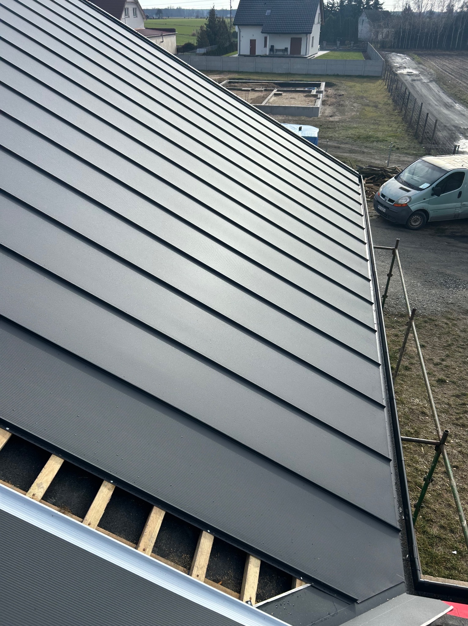 Nowe, grafitowe pokrycie dachowe z blachy na drewnianej konstrukcji, widoczne rynny i rusztowanie, w tle zabudowa jednorodzinna i zielony samochód dostawczy.