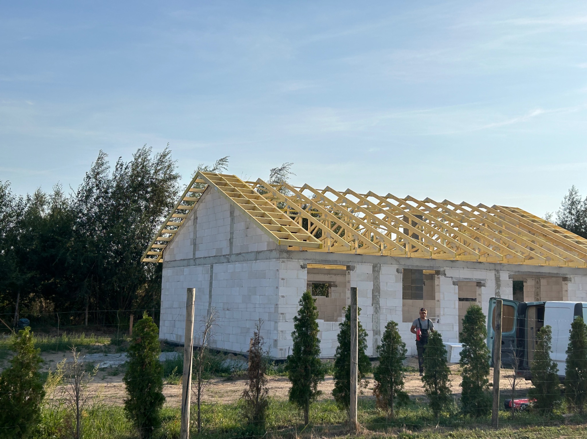 Wznoszenie konstrukcji dachu na budynku z białych bloczków, widoczna drewniana więźba dachowa, pracownik w tle i dostawczy samochód obok.