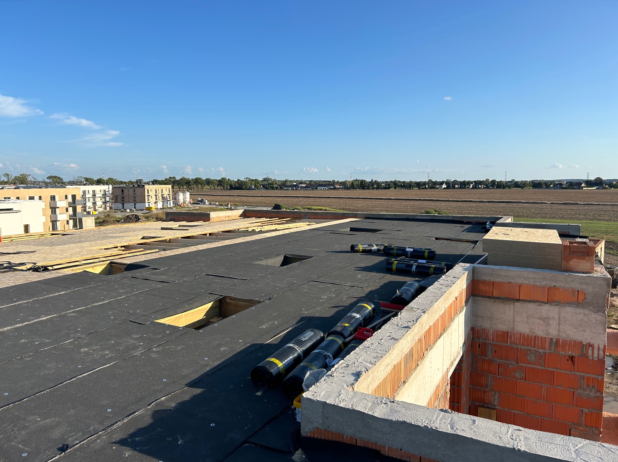 Montaż izolacji bitumicznej na dachu płaskim z widocznymi rolkami papy i fragmentami muru kominowego w tle krajobrazu.