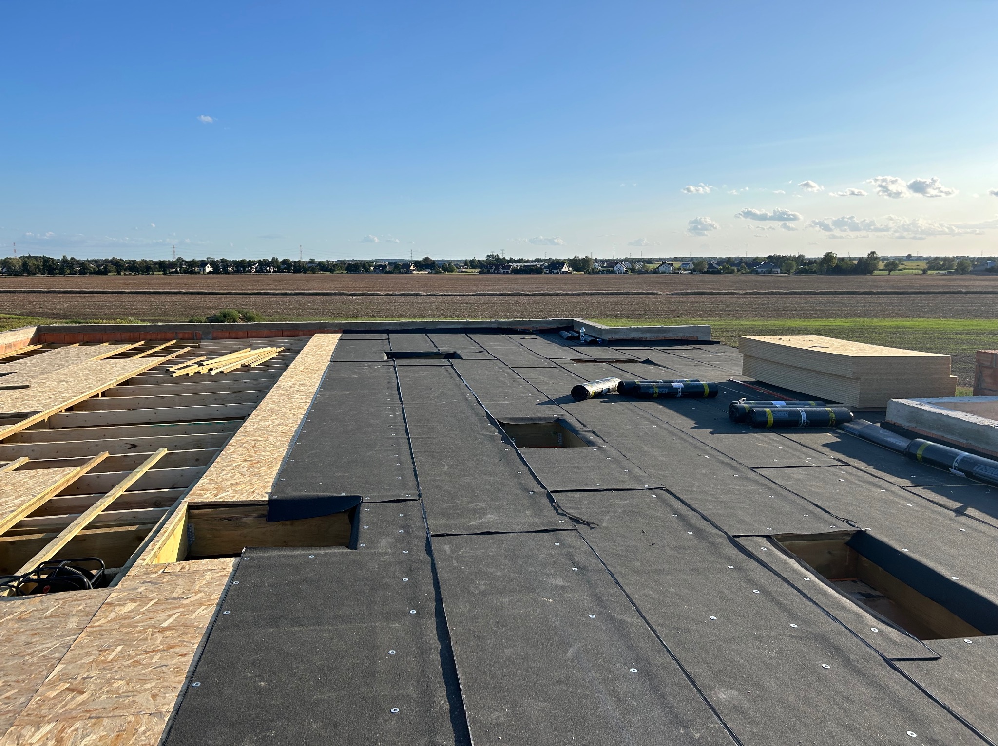 Płaski dach w trakcie budowy z widoczną konstrukcją drewnianą, płytami OSB i papą bitumiczną; widok na krajobraz polny z budynkami w oddali.