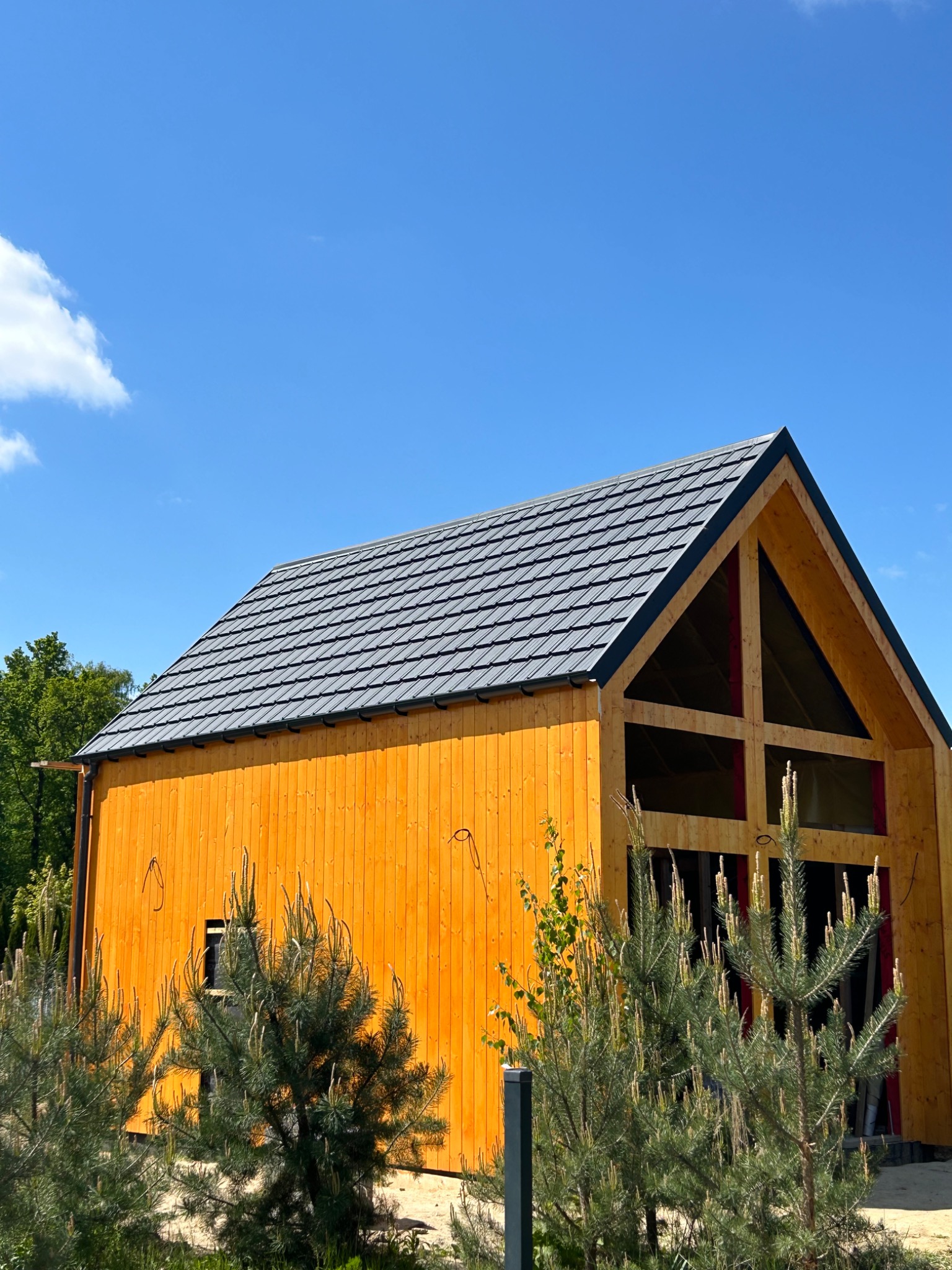 Nowoczesny dom z drewnianą elewacją i szarym dachem z dachówki, na tle błękitnego nieba. Widoczne detale wykończenia i roślinność w otoczeniu budynku.