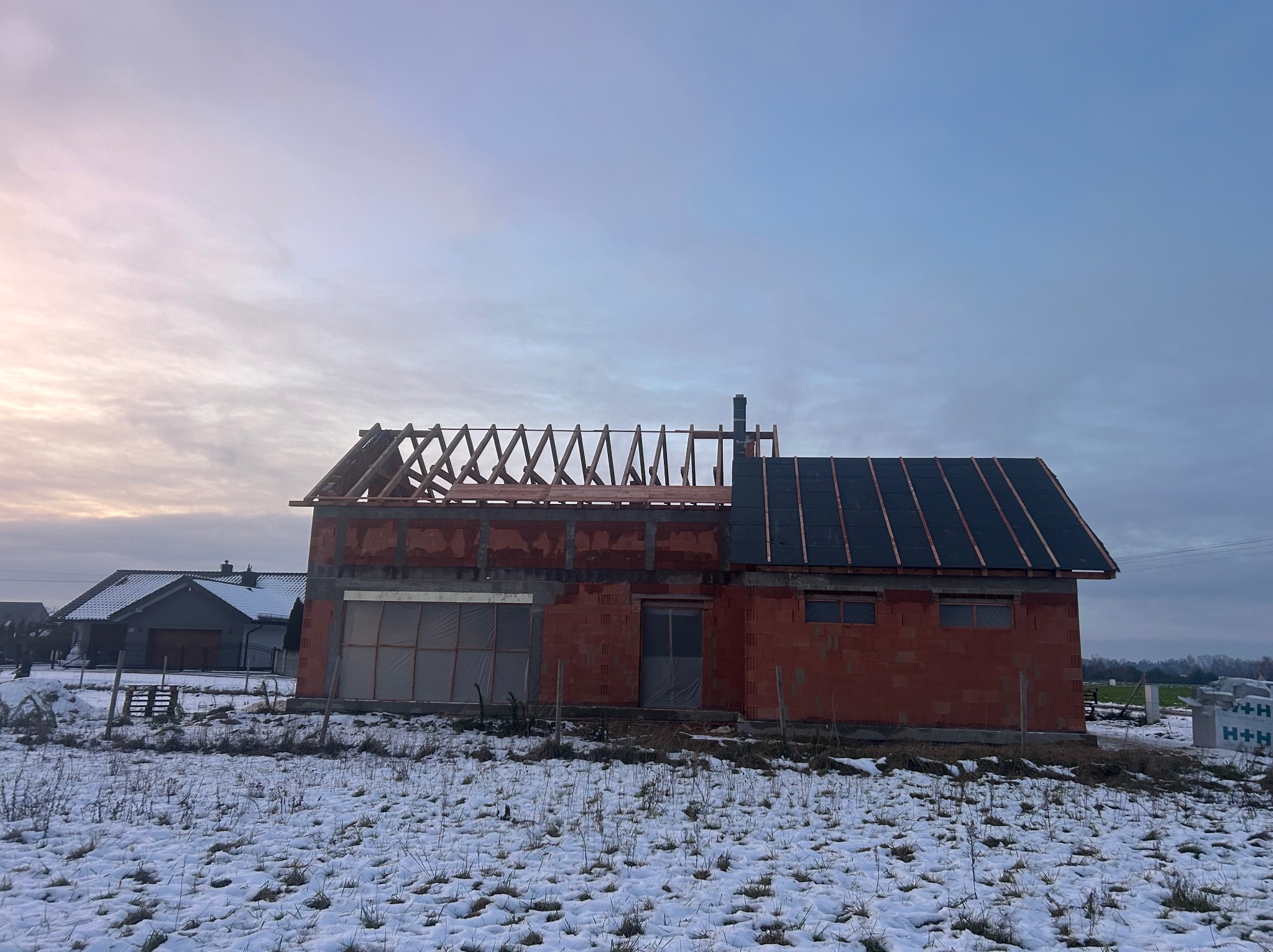 Dom w trakcie budowy: część dachu z czarną dachówką, część z widoczną więźbą. Zimowy krajobraz, ceglana konstrukcja, niebo z chmurami.