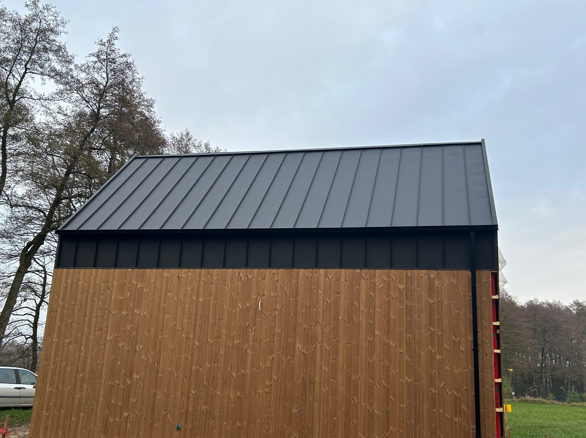 Nowoczesny dom z drewnianą elewacją i ciemnoszarym dachem z blachy na rąbek stojący. Widoczne drzewa w tle i fragment samochodu. Ujęcie z dołu.