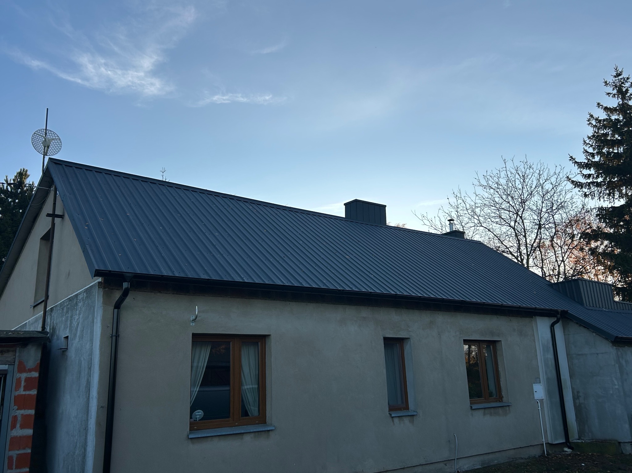 Dom jednorodzinny z nowym, ciemnym dachem z blachy trapezowej i zamontowanymi rynnami. Widoczne okna z drewnianą stolarką i fragment ściany z cegły.