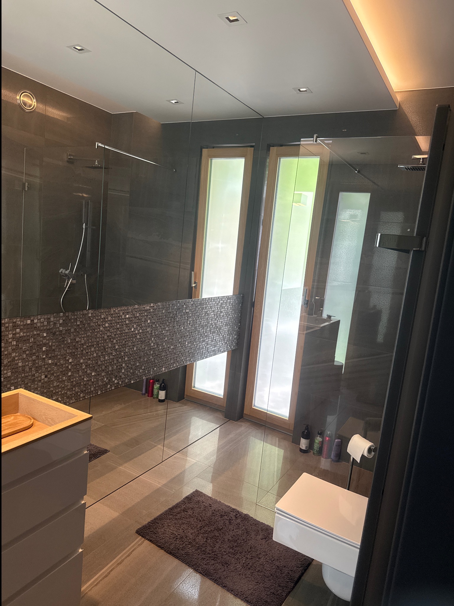 Nowoczesna łazienka z dużym lustrem, szarą mozaiką i drewnianymi oknami. Uwagę zwraca minimalistyczny design i dbałość o detale.