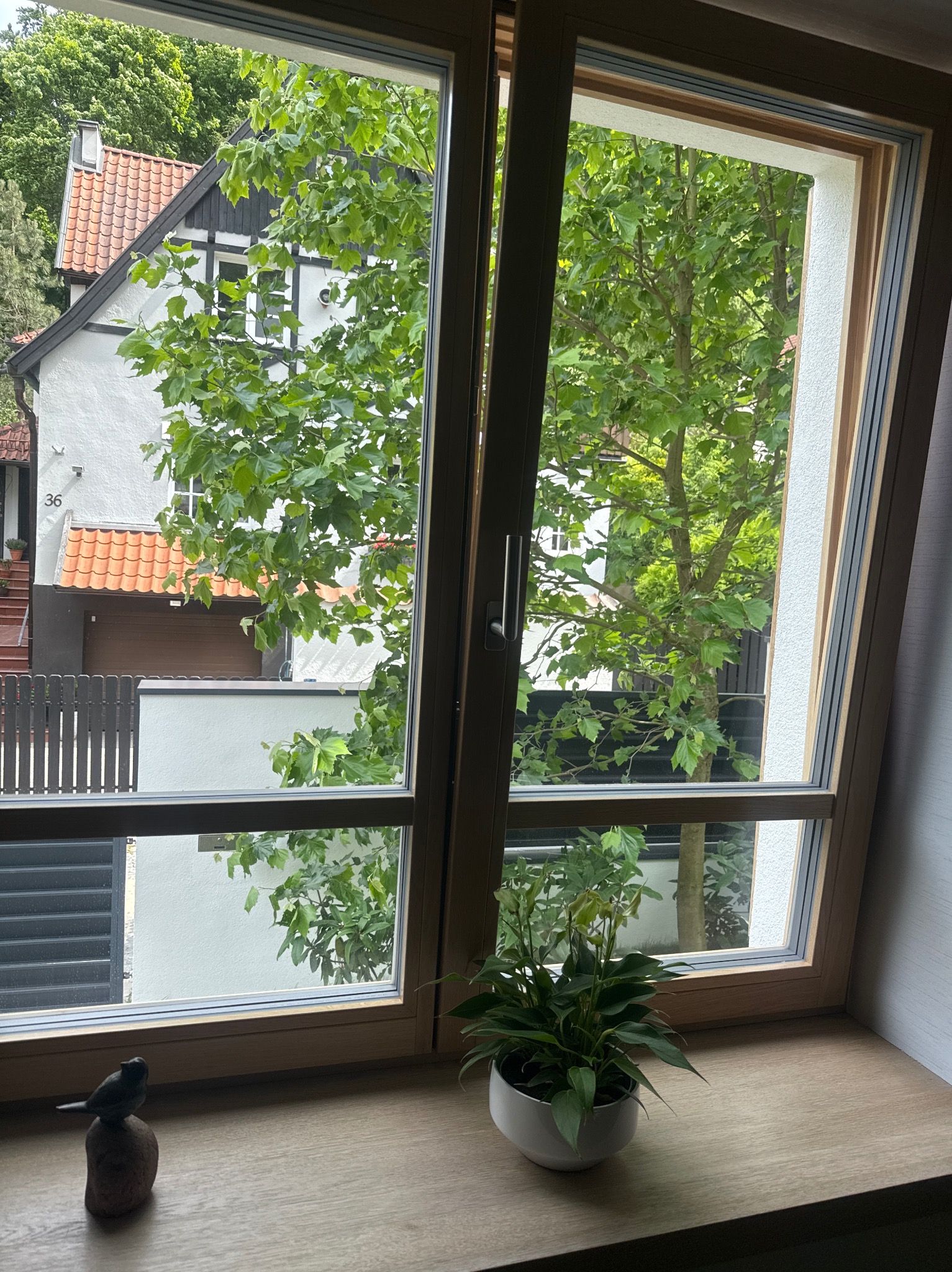 Otworzone okno z widokiem na dom z czerwonym dachem i drzewa. Na parapecie roślina w białej doniczce i mała figurka ptaka. Widok z bliska, naturalne światło.