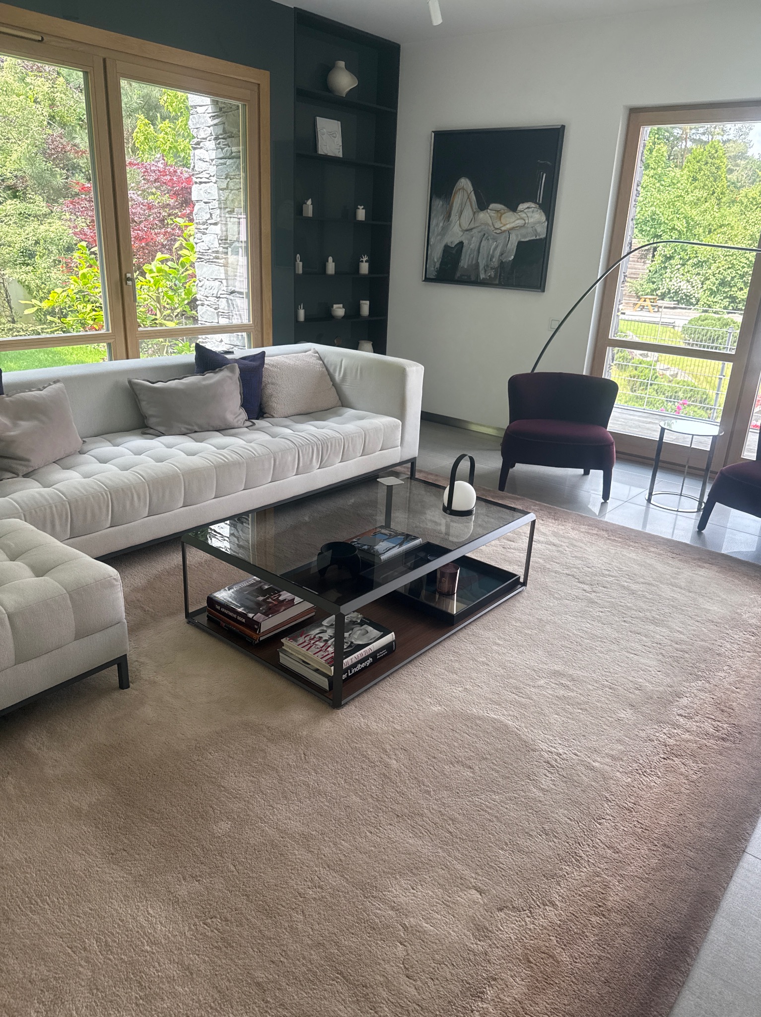 Jasny, przestronny salon z beżowym dywanem, białą sofą i stolikiem kawowym. Za oknem widok na zieleń ogrodu. Nowoczesne wnętrze z elementami dekoracyjnymi.