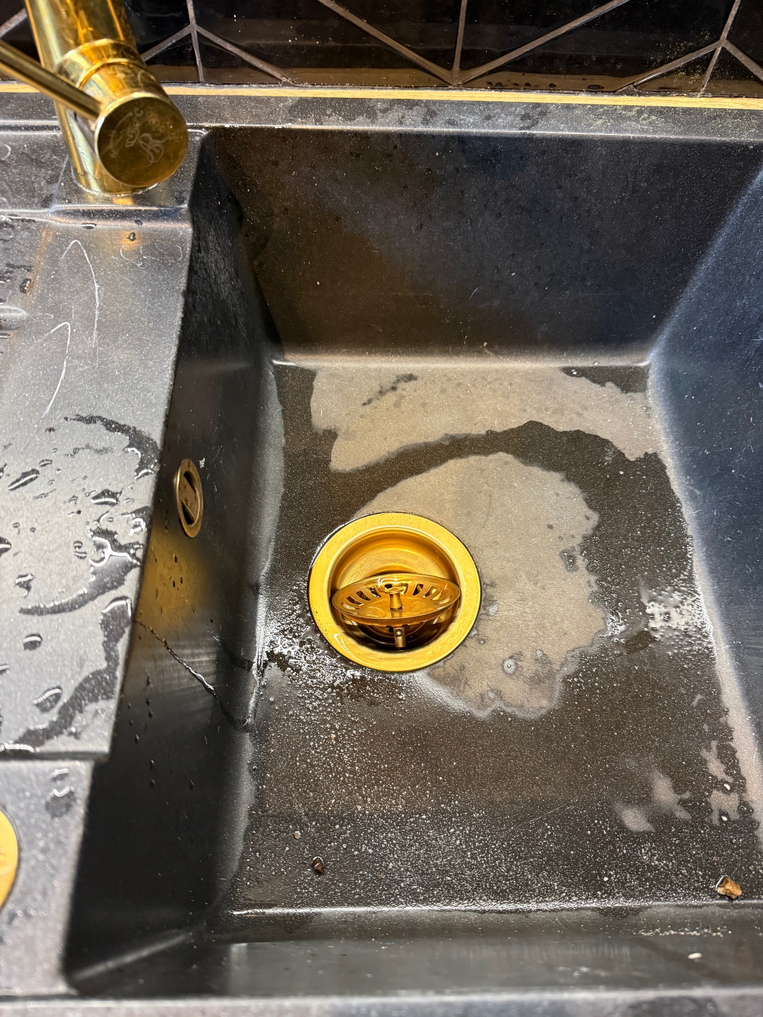 Czarny, granitowy zlew z złotą armaturą i odpływem. Widoczne ślady po umyciu, krople wody i delikatne zacieki. Ujęcie z góry, podkreślające fakturę zlewu.