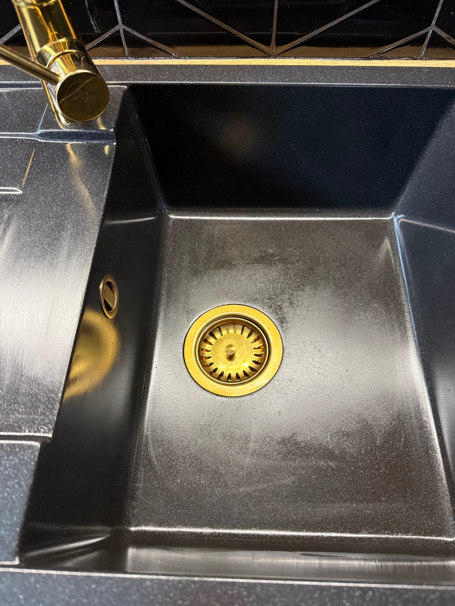 Błyszczący, czarny zlew granitowy z złotą armaturą i odpływem, po czyszczeniu. Widoczne refleksy światła na powierzchni. Górna część kadru z czarnymi płytkami.