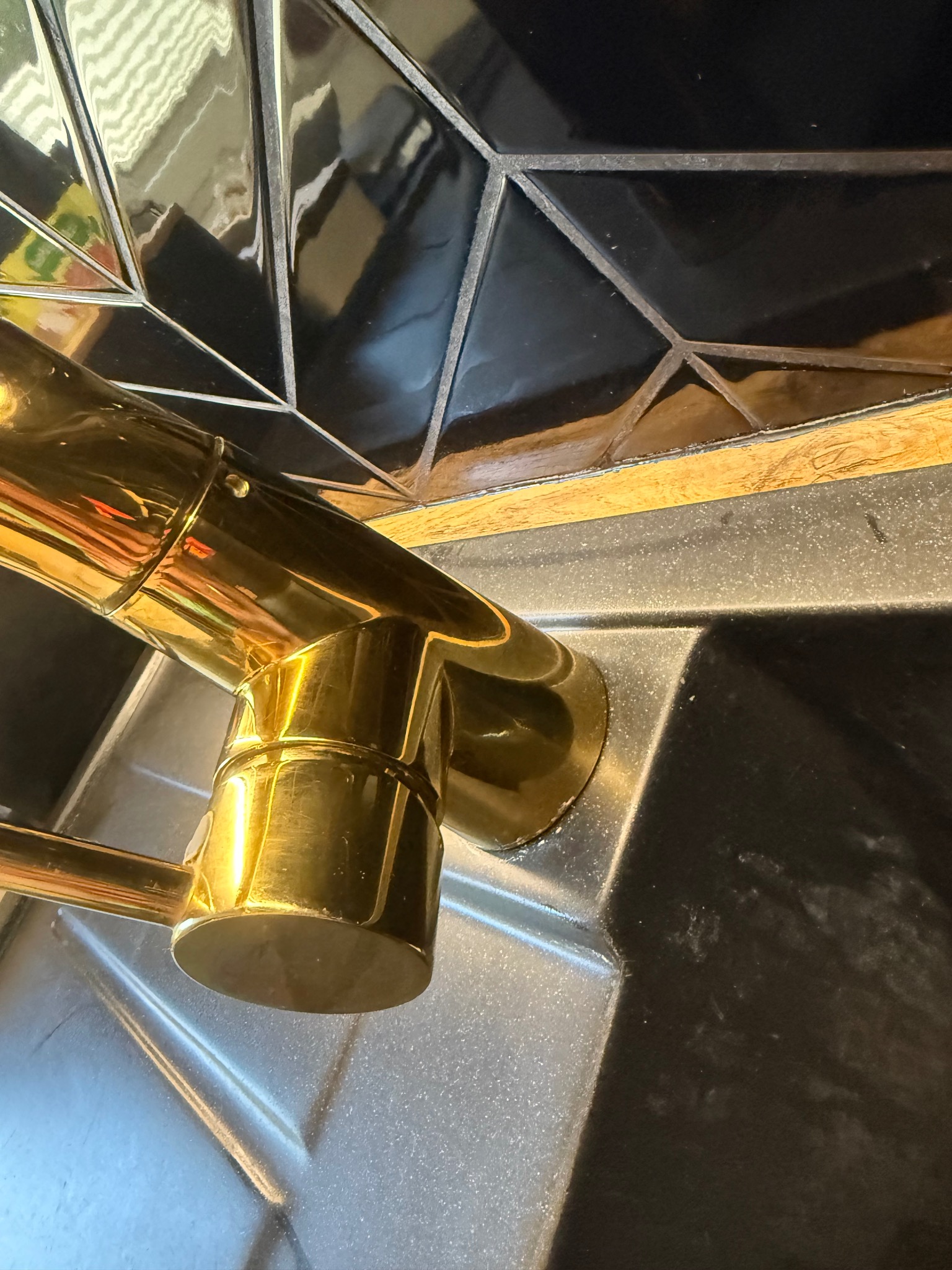 Błyszcząca, złota bateria umywalkowa na tle czarnej, geometrycznej mozaiki. Widoczne ślady po wodzie na umywalce. Ujęcie z bliska, podkreślające detale.