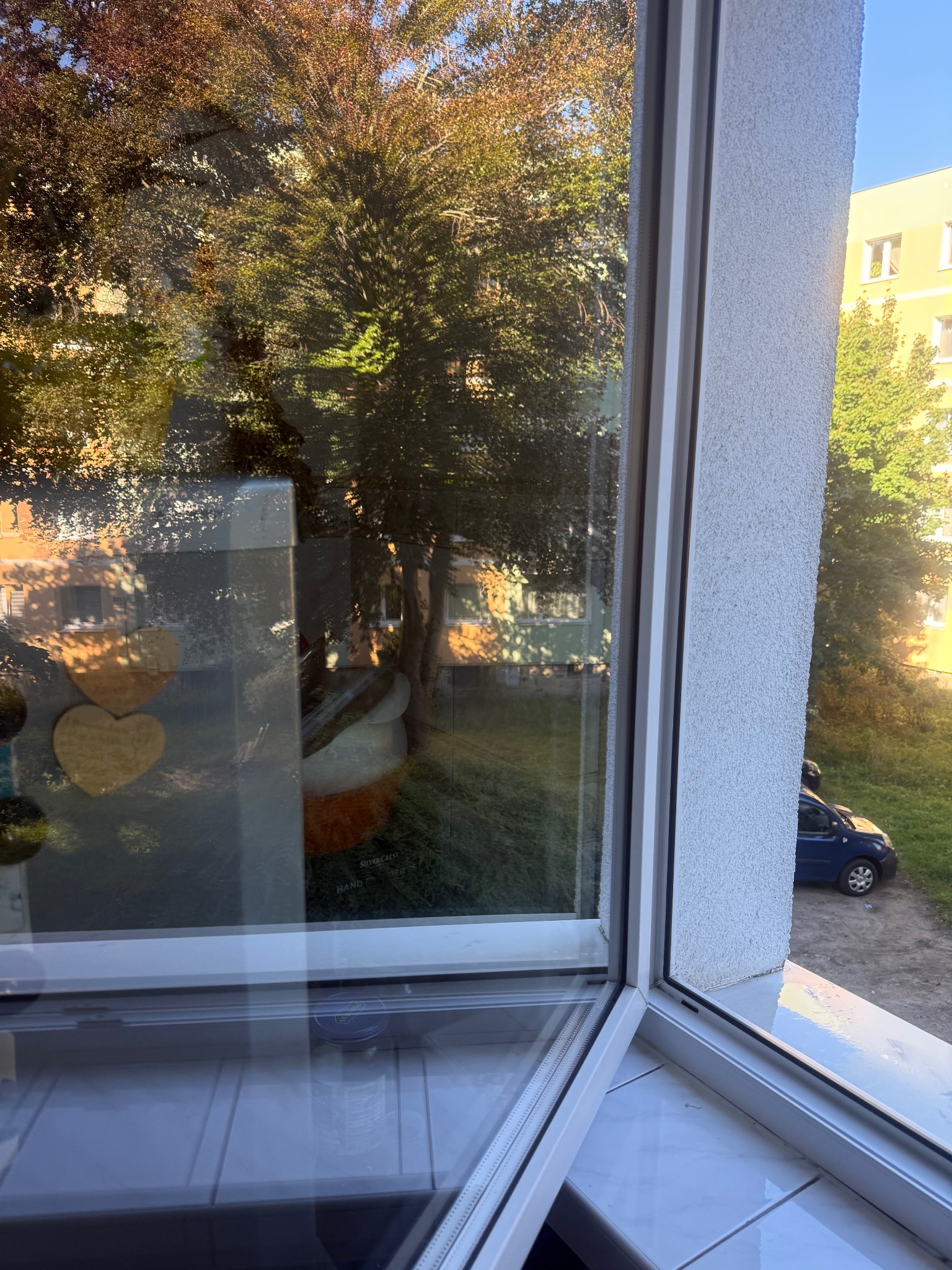 Otwarty biały oknem z widokiem na zewnątrz, gdzie widać drzewa, budynek i samochód. Na parapecie widać odbicie przedmiotów.