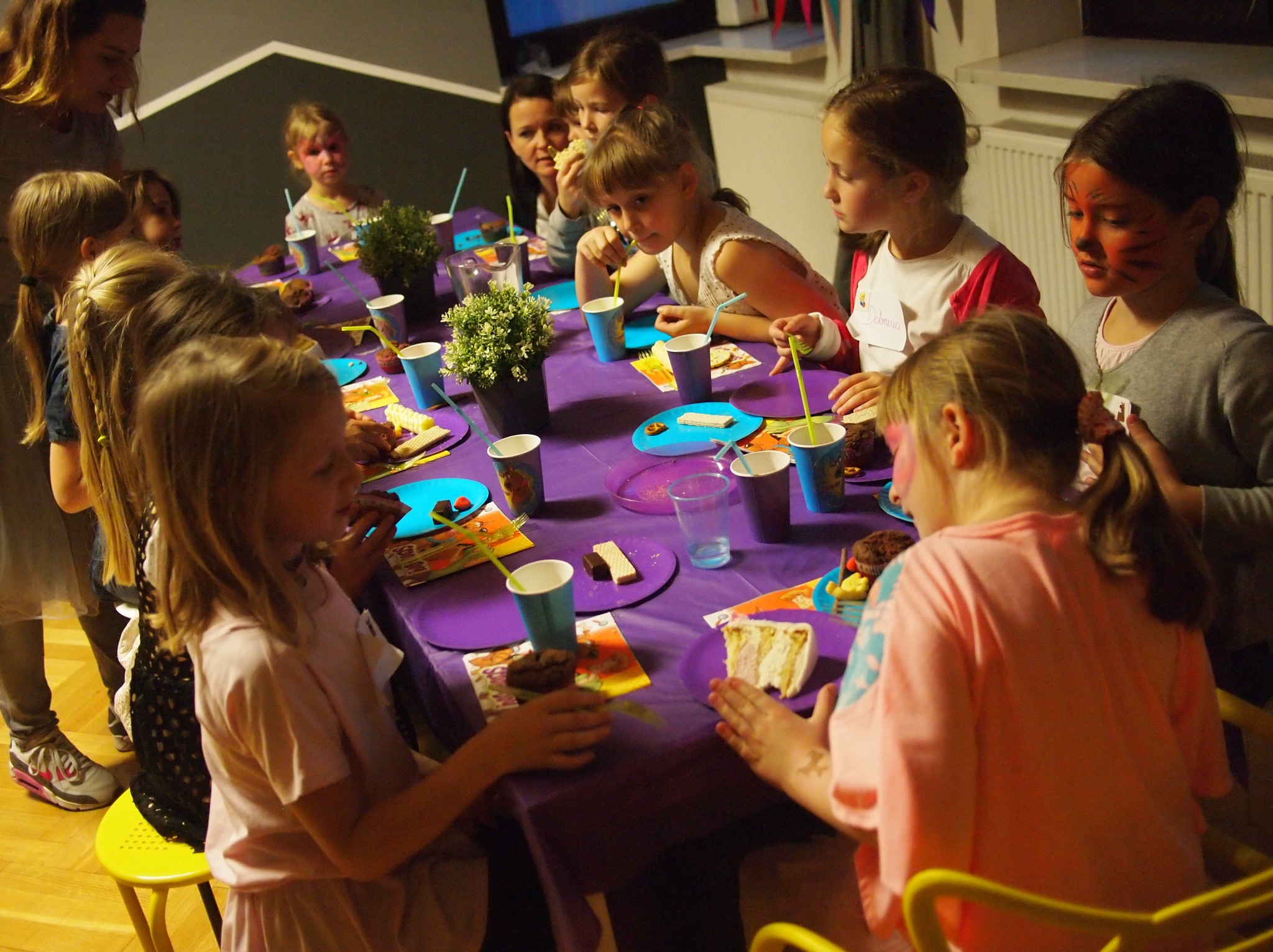 Urodziny dziecka: grupa dzieci siedzi przy fioletowym stole z przekąskami, napojami i dekoracjami. Niektóre dzieci mają pomalowane twarze.