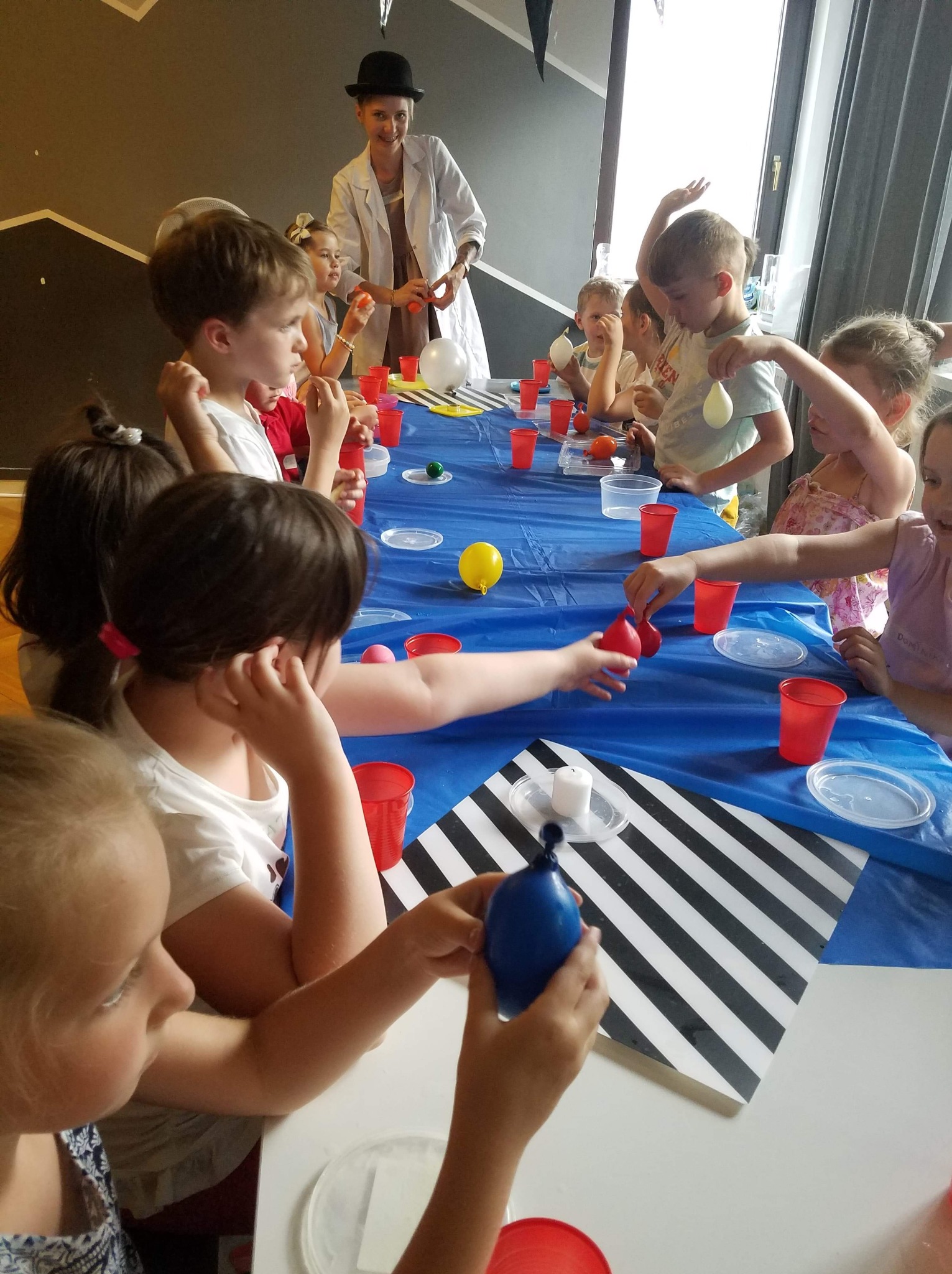 Dzieci przy stole z niebieskim obrusem, uczestniczące w eksperymentach naukowych prowadzonych przez kobietę w białym fartuchu i meloniku. Kolorowe balony i rekwizyty.