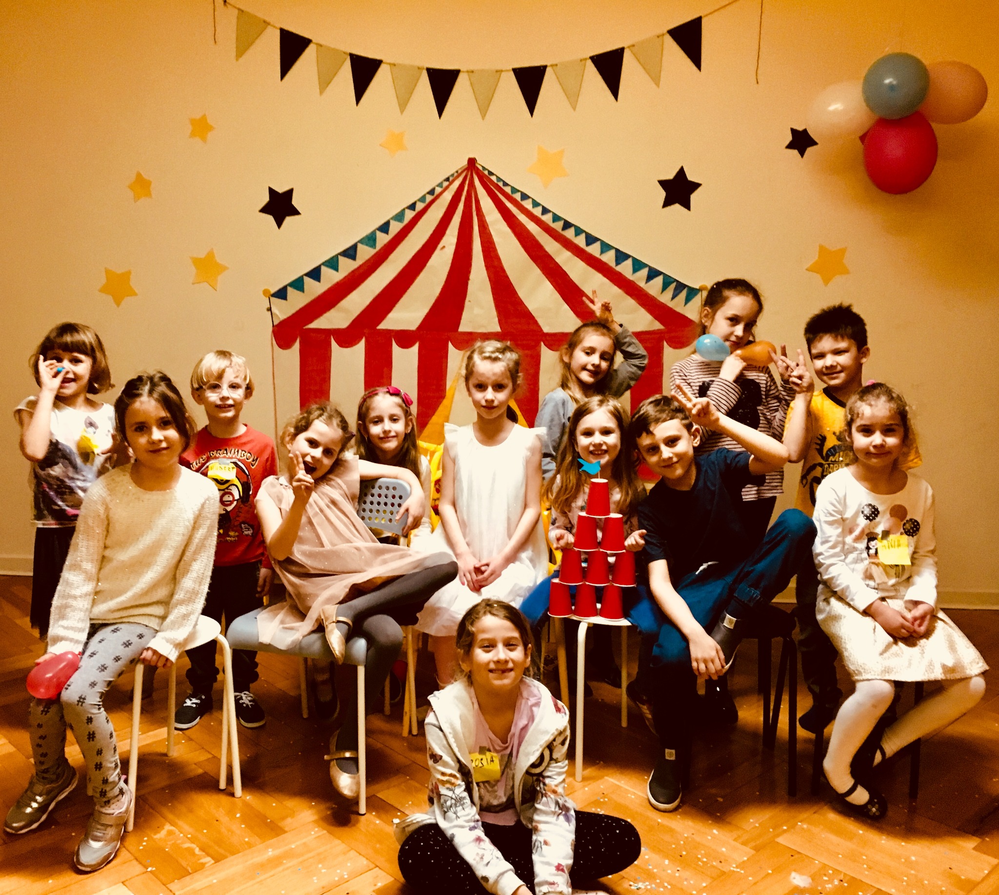 Grupowe zdjęcie dzieci na tle dekoracji cyrkowych, z piramidą z czerwonych kubków. Uśmiechnięte twarze, balony i konfetti tworzą radosną atmosferę.