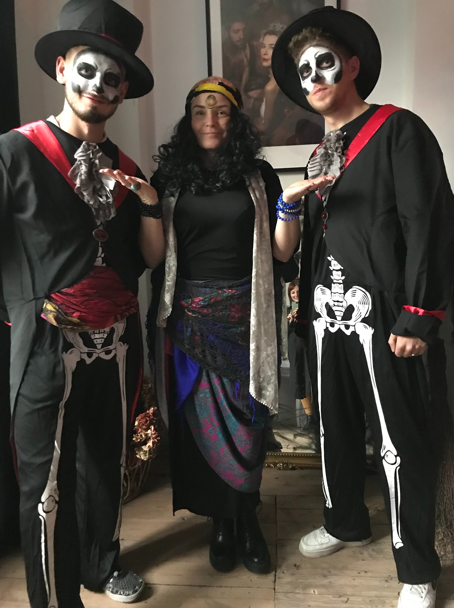 Trzy osoby w kostiumach na Halloween: dwaj mężczyźni w strojach szkieletów i kapeluszach oraz kobieta w stroju cyganki z chustą na głowie i kolorową spódnicą.