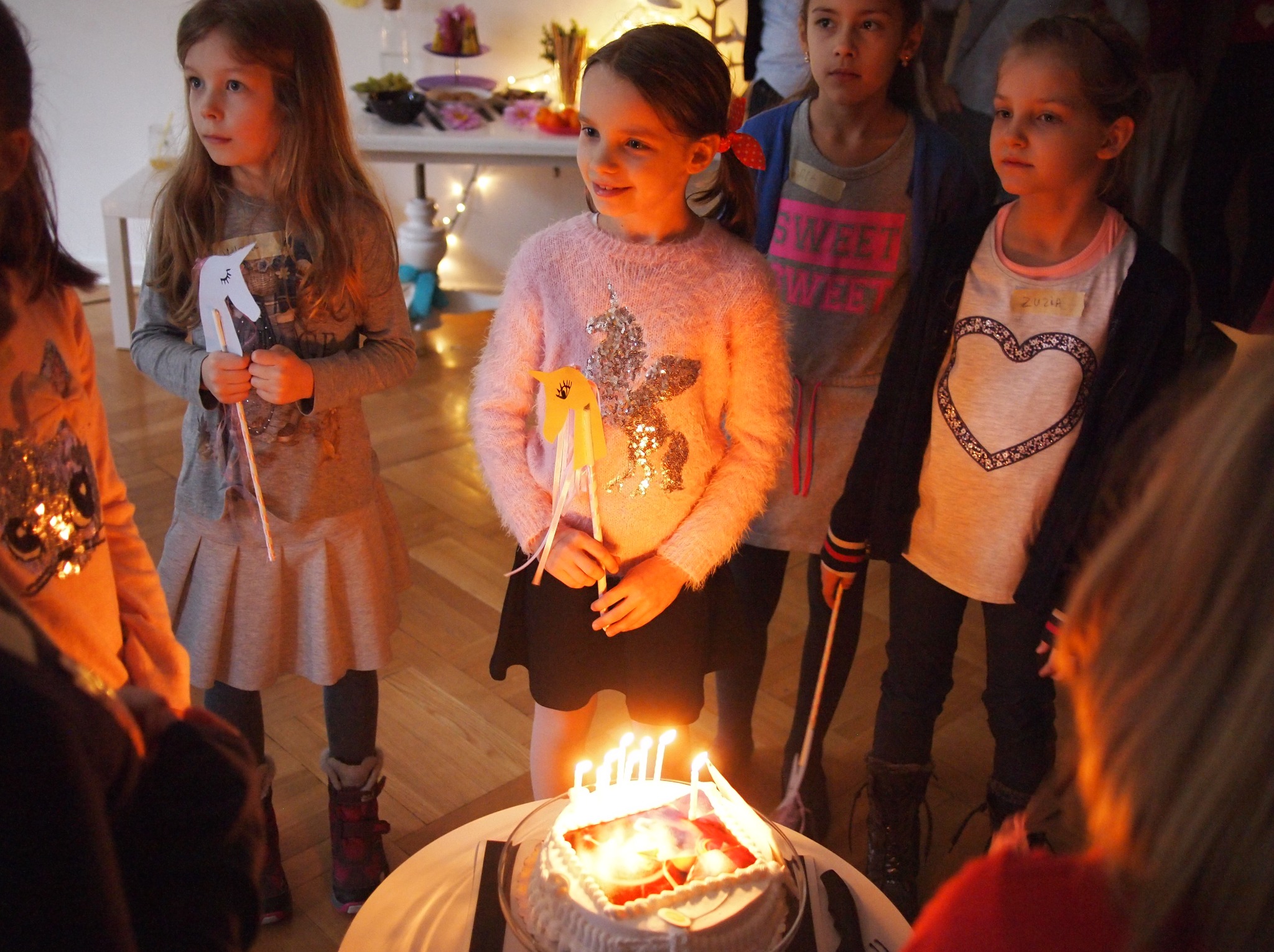 Uśmiechnięte dziewczynki z papierowymi jednorożcami wokół tortu urodzinowego ze świeczkami, w tle stół z przekąskami. Impreza urodzinowa.