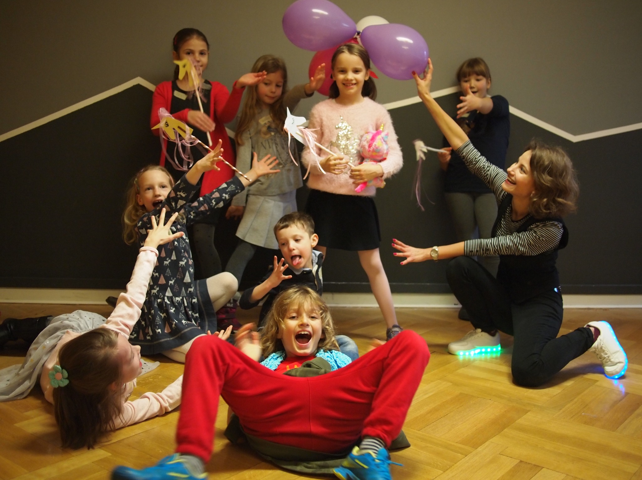 Radosna grupa dzieci z balonami i rekwizytami na imprezie, prowadzona przez animatorkę w podświetlanych butach. Dynamiczne ujęcie z perspektywy podłogi.