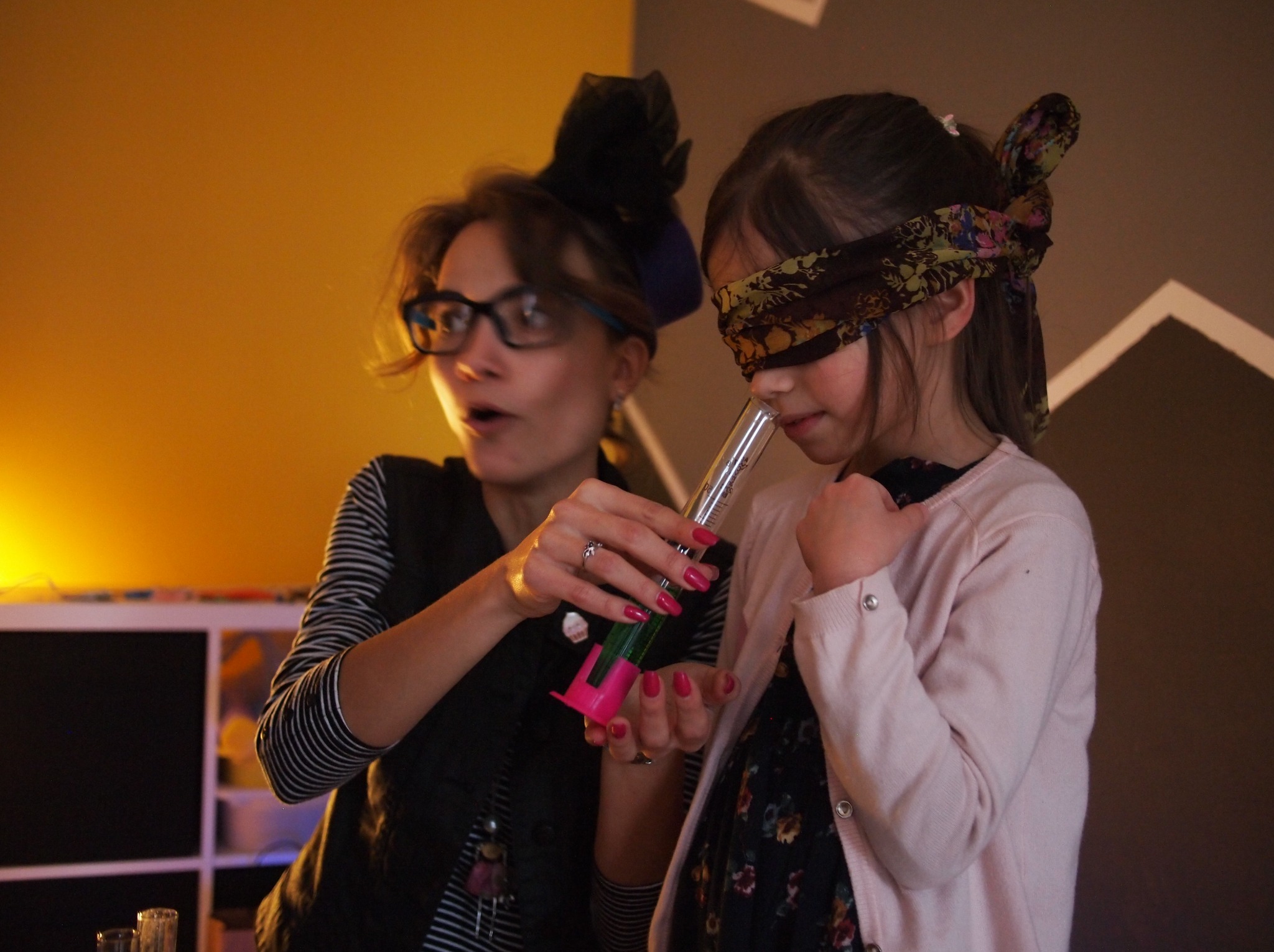 Animator prezentuje eksperyment chemiczny z dużą probówką dziewczynce z zasłoniętymi oczami. Nietypowe nakrycie głowy animatorki. Żółte oświetlenie.