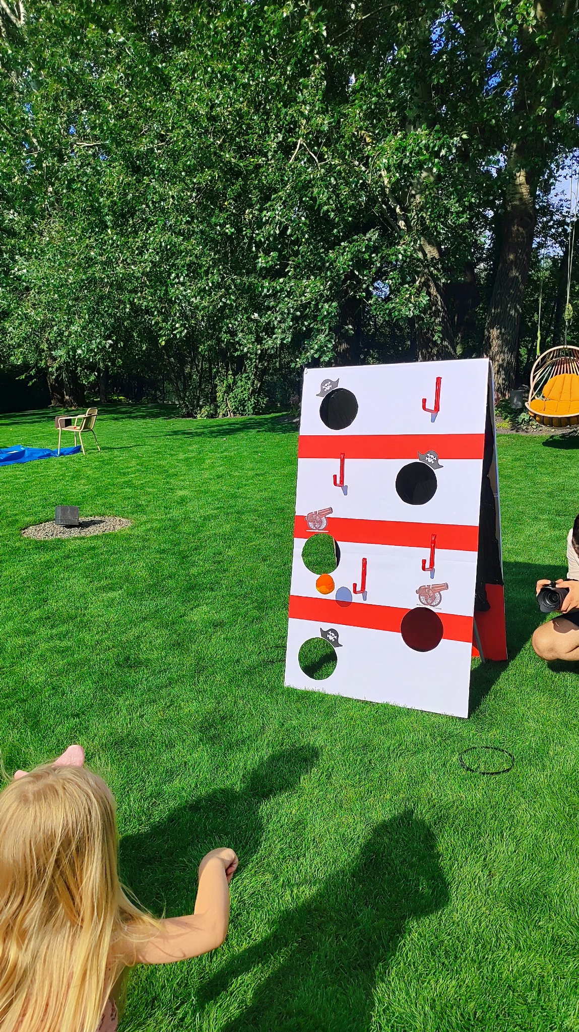 Dziewczynka rzuca piłką do pirackiej gry z otworami na trawie, w tle drzewa i huśtawka. Zabawa na świeżym powietrzu, impreza tematyczna.