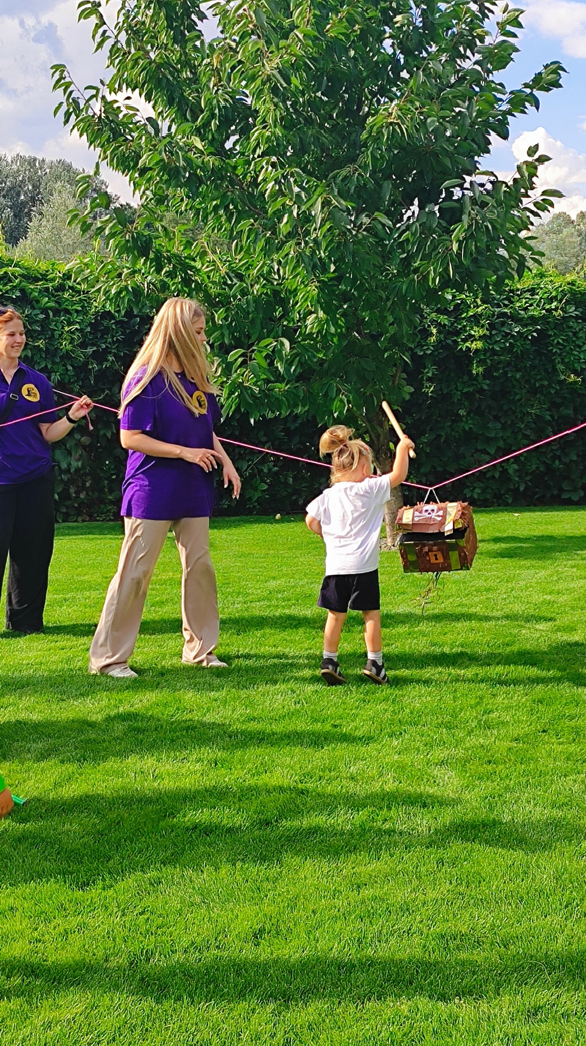 Dziewczynka uderza kijem w piniatę w kształcie skrzyni skarbów, trzymaną przez dwie osoby na zielonym trawniku podczas imprezy plenerowej.