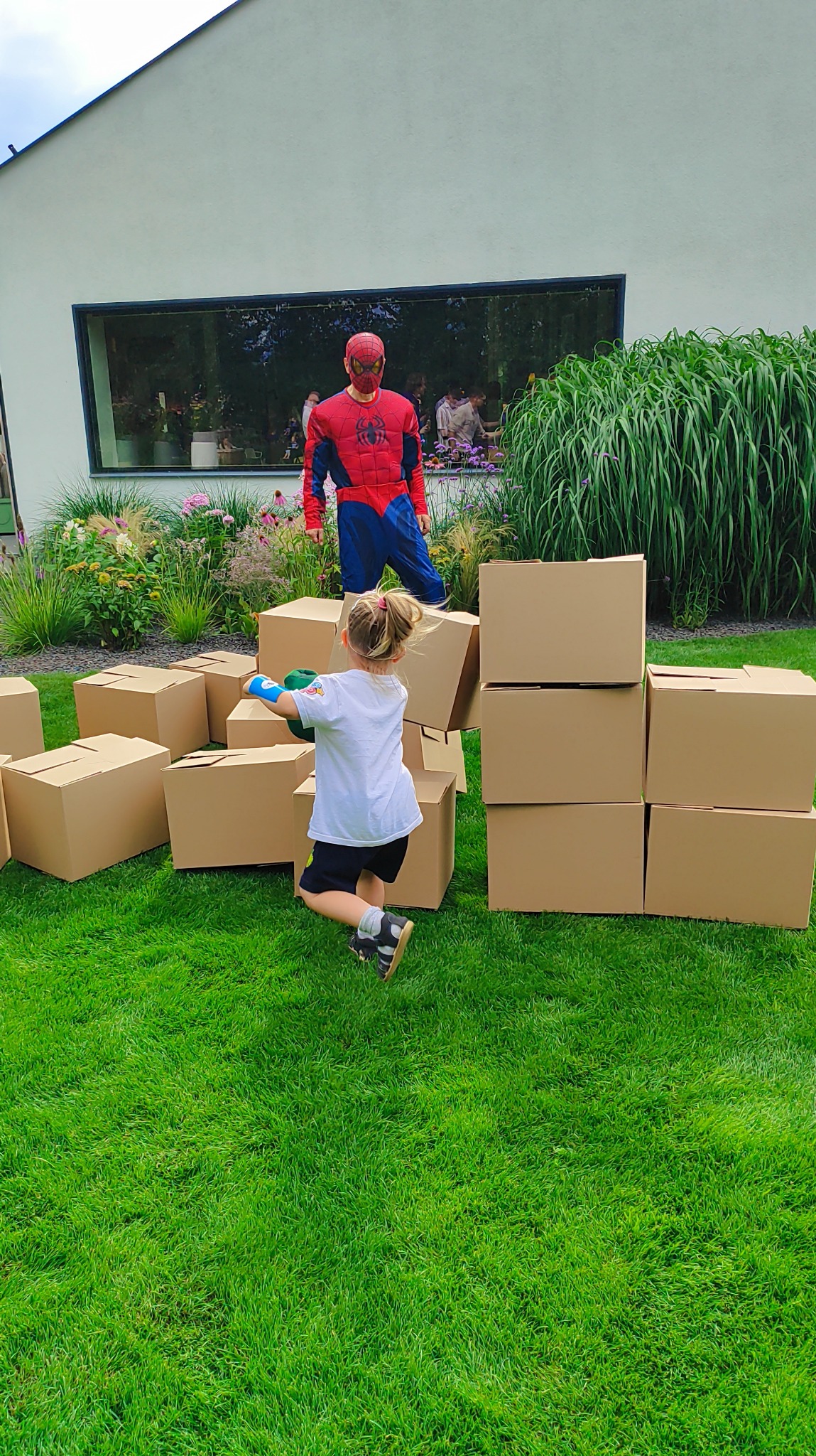 Dziewczynka bawi się kartonami na trawie, w tle postać w kostiumie Spider-Mana. Zabawa na świeżym powietrzu, animacje dla dzieci, impreza plenerowa.