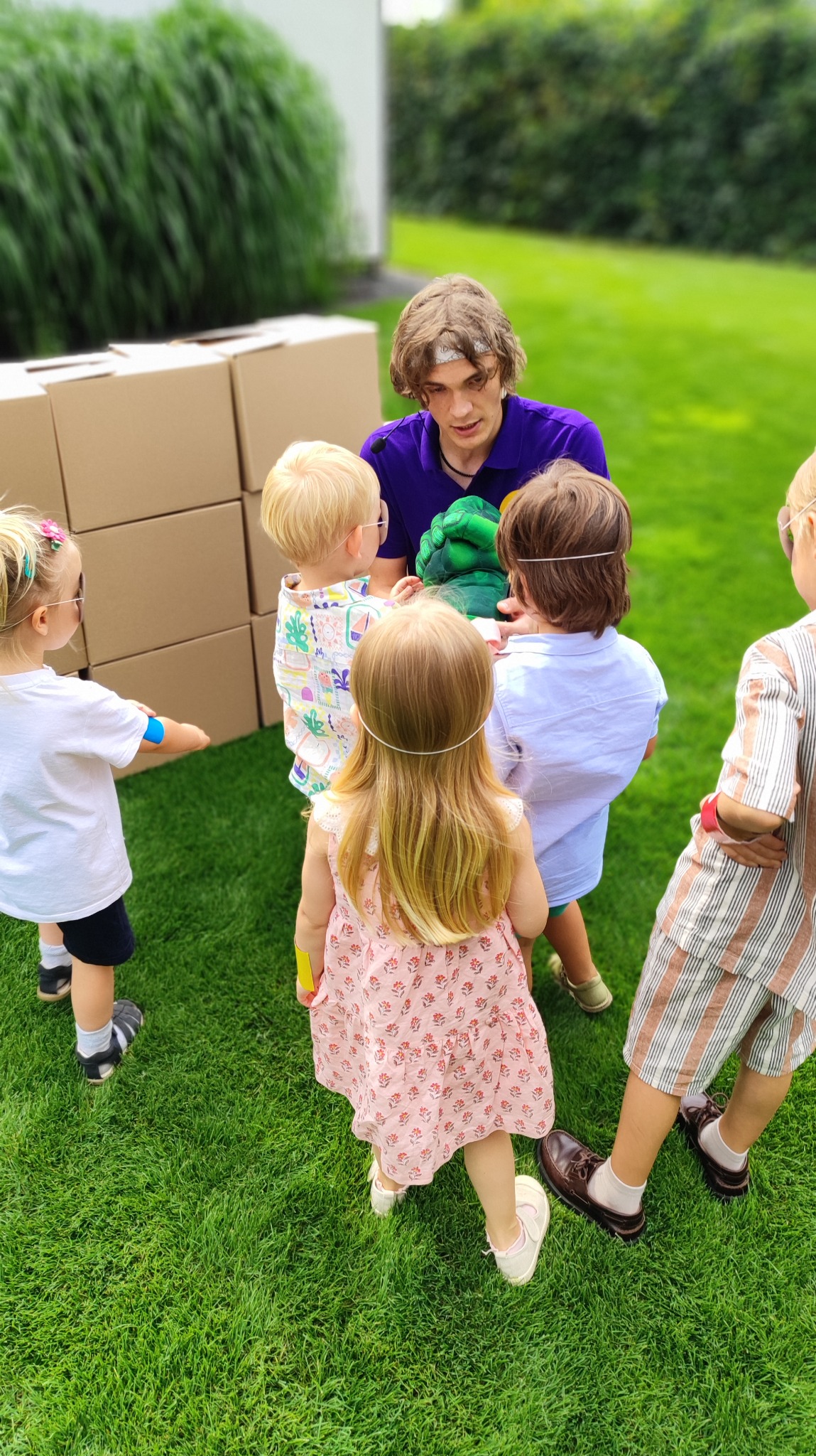 Animator w fioletowej koszulce z zieloną pacynką otoczony przez grupę dzieci na trawie, z kartonami w tle. Ujęcie z góry, plener.