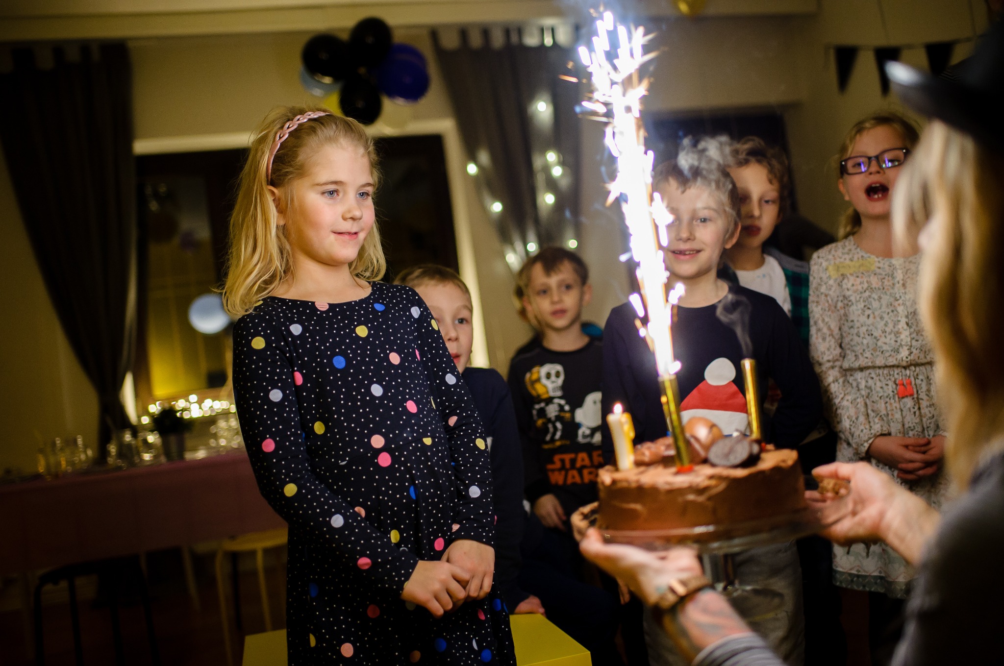 Uśmiechnięta dziewczynka w sukience w kropki, w tle dzieci i tort z fontanną iskier na przyjęciu urodzinowym, trzymany przez osobę z tatuażami na rękach.