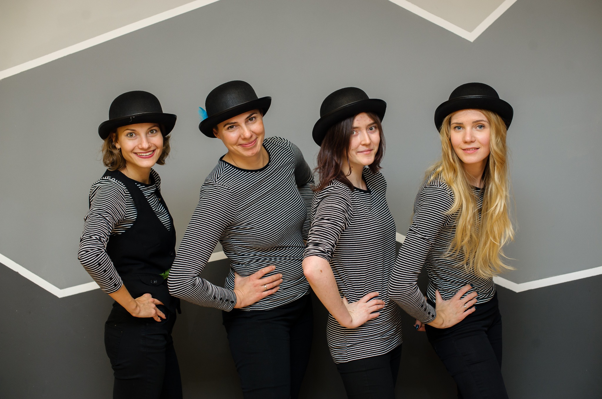 Cztery uśmiechnięte kobiety w czarnych melonikach i pasiakach pozują na tle szaro-białej ściany. Jedna z nich ma niebieskie piórko w kapeluszu.