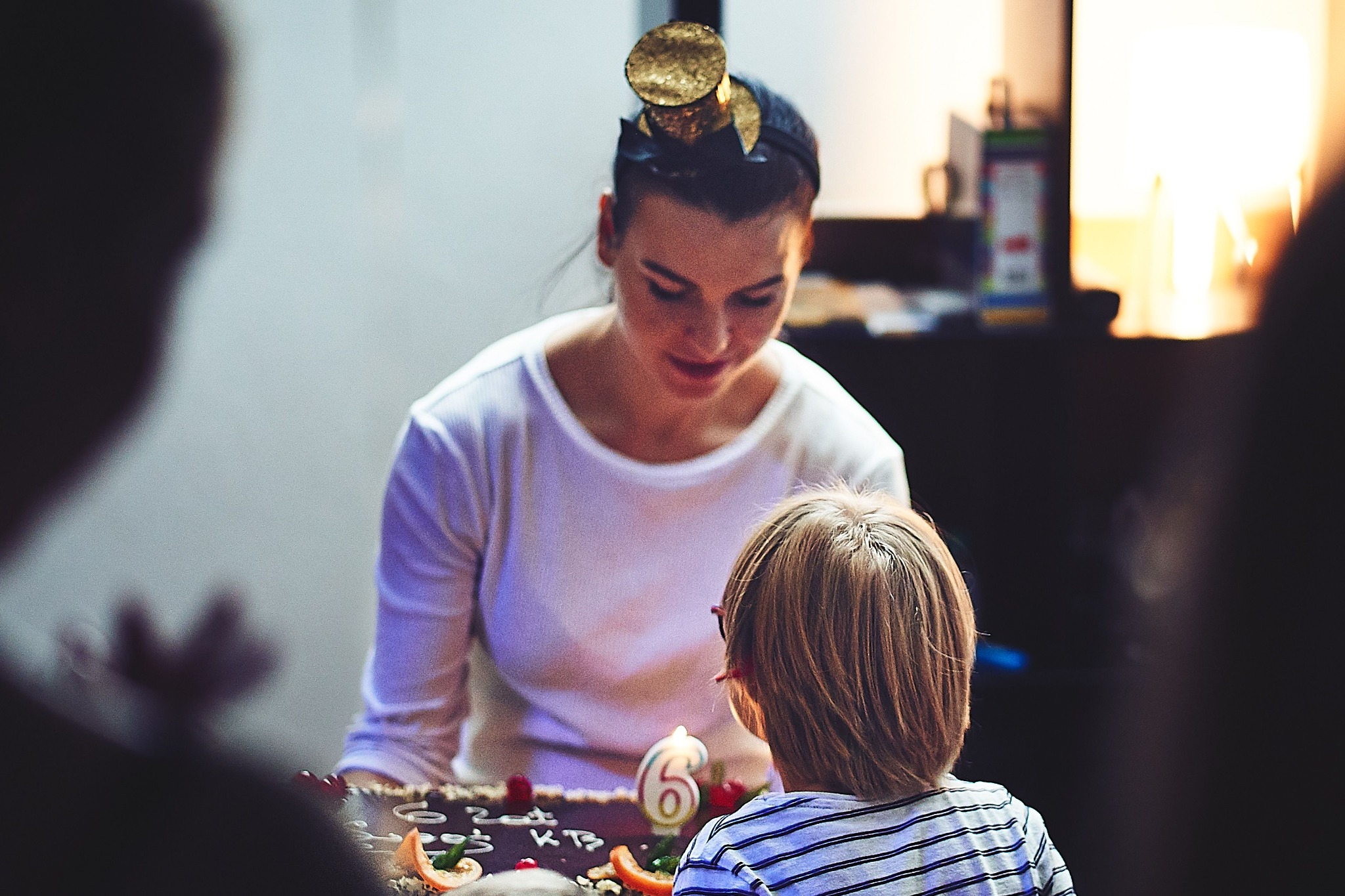 Animator w złotym nakryciu głowy, pochylona nad tortem urodzinowym z płonącą świeczką w kształcie cyfry 6, wpatrująca się w dziecko w paski z tyłu.