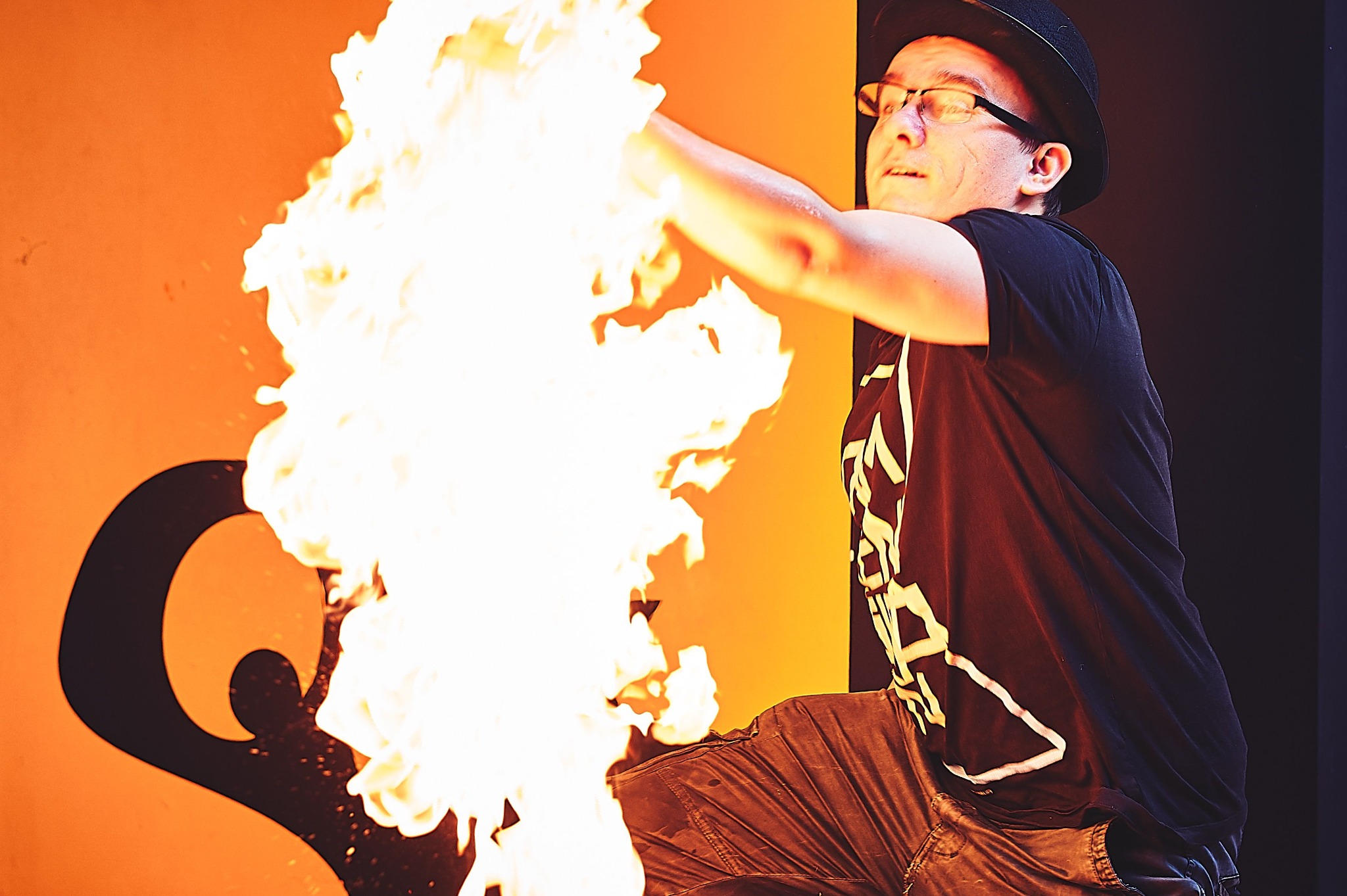 Mężczyzna w kapeluszu i okularach prezentuje pokaz z ogniem na tle pomarańczowej ściany z czarnym logo. Dynamiczne ujęcie, jasne płomienie, ciemna odzież.