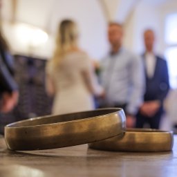 Zatrzymane w Czasie - Dwa złote obrączki ślubne leżące na drewnianym stole, w tle rozmyta para młoda i goście podczas ceremonii.