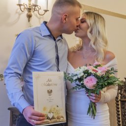 Zatrzymane w Czasie - Pocałunek pary młodej trzymającej akt małżeństwa i bukiet w jasnym wnętrzu z ozdobnym żyrandolem.