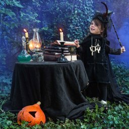 Zatrzymane w Czasie - Uśmiechnięta dziewczynka w kostiumie czarownicy z różdżką, pozuje obok stolika z dynią, świecami, książkami i eliksirami w scenerii leśnej z mgłą.