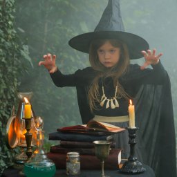 Zatrzymane w Czasie - Dziewczynka w kostiumie czarownicy, z kapeluszem i naszyjnikiem z kości, stoi przy stole z eliksirami i świecami, w tle zarośnięty ogród.