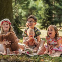 Zatrzymane w Czasie - Czterech roześmianych dzieci siedzi na kocu piknikowym w lesie, jedno trzyma jabłko, drugie pluszowego misia, a w tle widoczny jest wiklinowy koszyk.