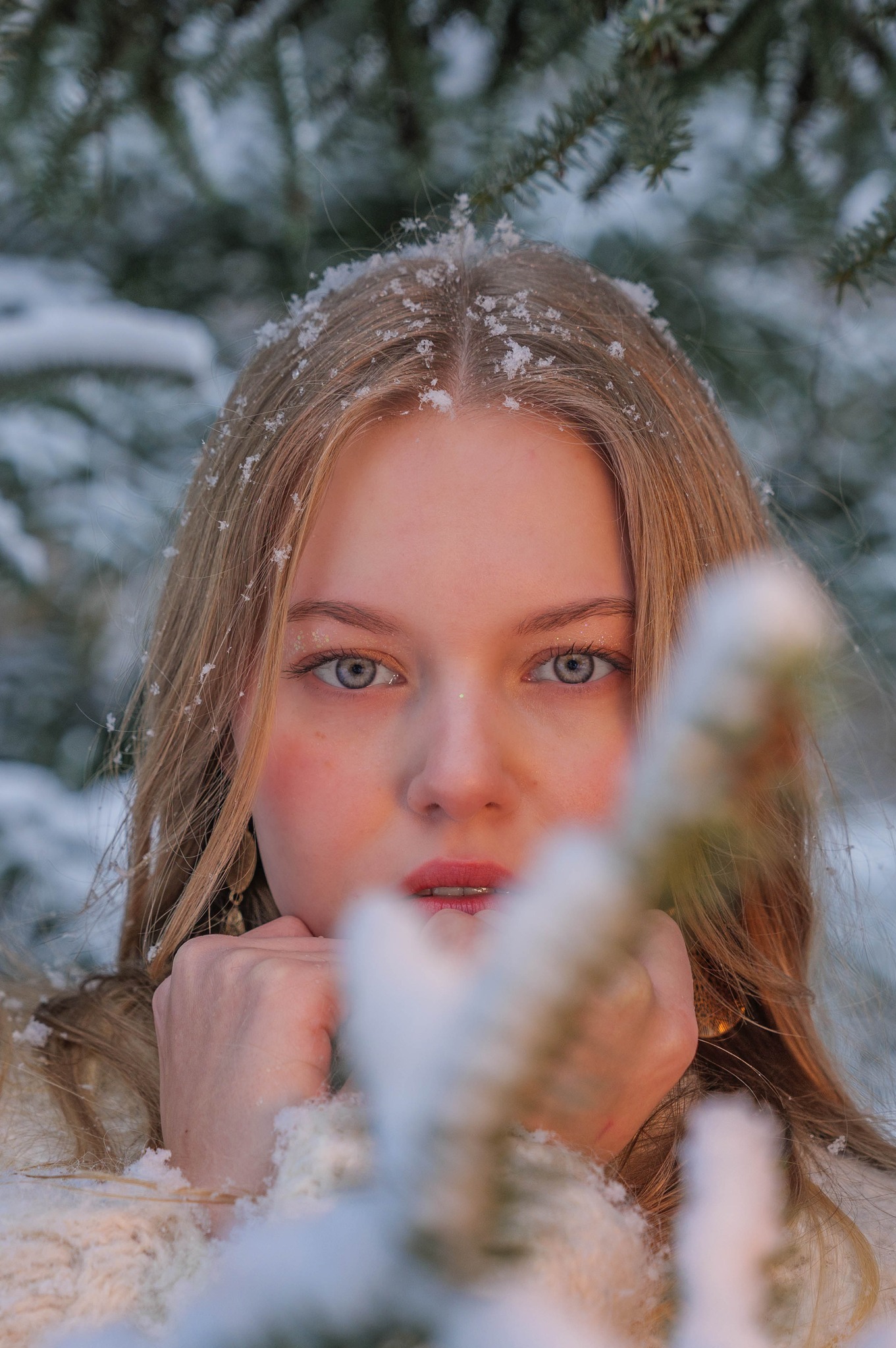 Portret młodej kobiety z blond włosami, oprószonej śniegiem, z delikatnym makijażem i spojrzeniem skierowanym wprost, na tle zimowego krajobrazu.