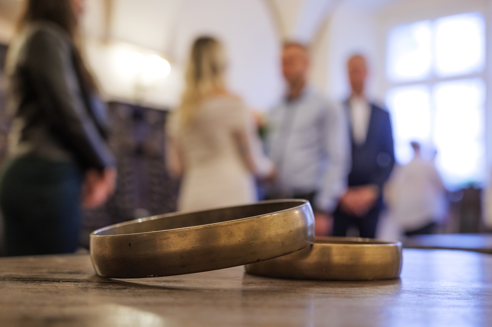 Dwa złote obrączki ślubne leżące na drewnianym stole, w tle rozmyta para młoda i goście podczas ceremonii.